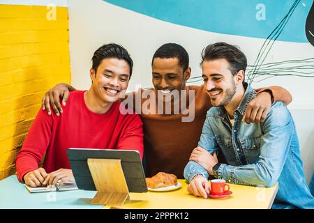 Guy friends sitting at a table in a café during breakfast, watching a funny video on their tablet with amusement. Moment of fun and relaxation. Stock Photo