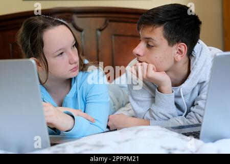 teenage pimples on face boy and girl friends are sitting at computer we are lying on stomachs learning lessons from boy on cheek a lot of teenage rash girl is looking carefully solve problem Stock Photo