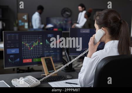 Rear view of young broker talking on the phone with customer while analyzing graphs on monitors sitting at her workplace Stock Photo