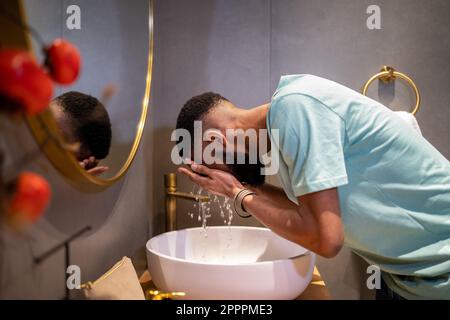 African American millennial guy cleaning skin in morning, washing face with water in bathroom Stock Photo