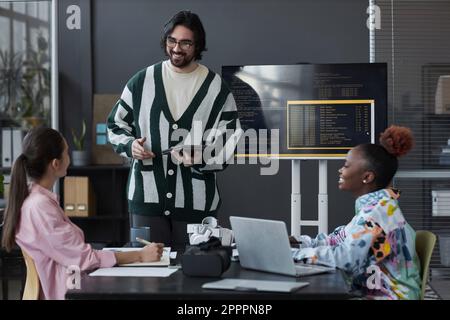 Young developer standing near the screen with codes and presenting his report to colleagues during teamwork in office Stock Photo