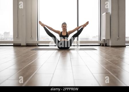 Front view of beautiful fitness model in black sportswear doing stretching while sitting on floor of white capacious studio. Caucasian dark haired lady balancing on yoga mat during everyday training. Stock Photo