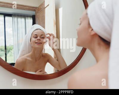 A beauty woman stands in front of a mirror after a shower in a towel on her head looks at her reflection and does a facial massage applies a day cream Stock Photo