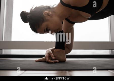 Fitness dark haired woman wearing black crop top and leggings stretching legs on yoga mat during morning workout. Charming caucasian brunette enjoying active time spending at spacious studio. Stock Photo