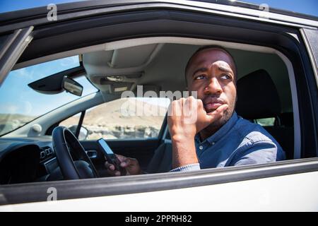 An African businessman sitting in his car with a thoughtful expression while using his smartphone. Stock Photo