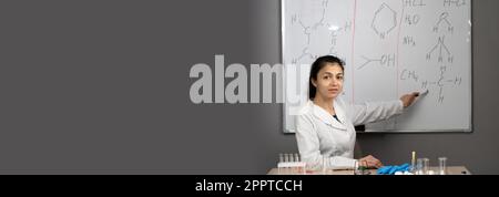 A Chemistry teacher sitting at desk and pointing on blackboard, teaching in a classroom setting Stock Photo