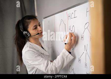 Hispanic female school teacher working from home, teaching chemistry formulas subject to students online. Woman pointing at whiteboard, talking with Stock Photo