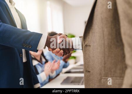 Close-up of two businessmen shaking hands in an office setting with applauding colleagues in the background, faces not visible. Stock Photo