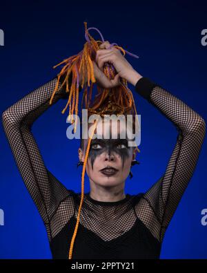 Informal young female with colored braids hairdo and spooky black stage makeup painted on face. Front view, studio shot on blue background. Part of ph Stock Photo