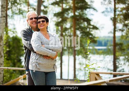 Mature couple standing on porch and hugging. They are smiling and looking at the beautiful view - forest and lake. Happy senior couple embracing Stock Photo