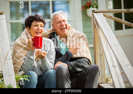 Mature couple standing on porch and drinking hot coffee from mug. They are smiling and laughing, wrapped in a blanket. Happy senior couple embracing Stock Photo