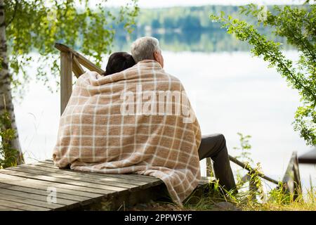 Mature couple sitting on wooden steps and hugging. They are wrapped in blanket, smiling and looking at beautiful view - forest and lake. Happy senior Stock Photo