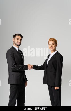 successful business partners in black blazers shaking hands and looking at camera isolated on grey,stock image Stock Photo