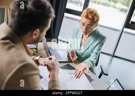 frowning businesswoman in eyeglasses pointing at analytics near blurred colleague in office,stock image Stock Photo
