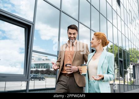 cheerful business colleagues with takeaway drinks talking while walking along modern building on urban street,stock image Stock Photo