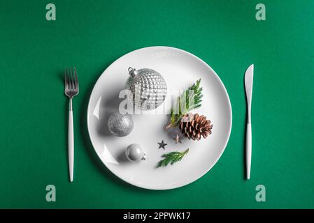 Creative Christmas Table Setting.  Festive ornaments and decor on white plate on green background, top view, copy space. Christmas dinner, party desig Stock Photo