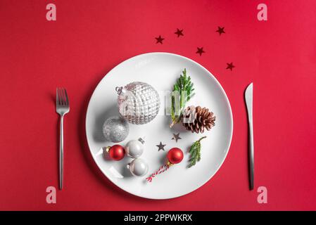 Beautiful Christmas Table Setting.  Festive silver baubles and pine cone on white plate on red background, creative flat lay. Christmas dinner, party Stock Photo