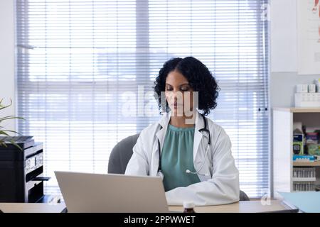 Biracial female doctor sitting at desk and using laptop at doctor's office Stock Photo