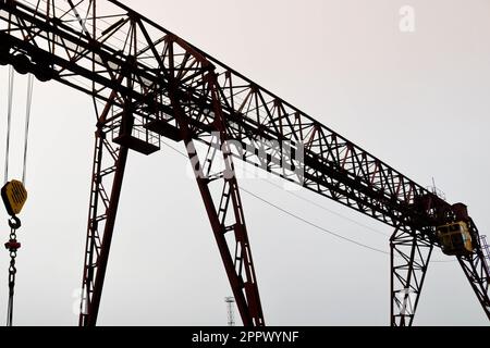 Industrial iron large metal gantry crane with a hook mounted on the supports for lifting and carrying heavy cargo, moving along the rails at the facto Stock Photo