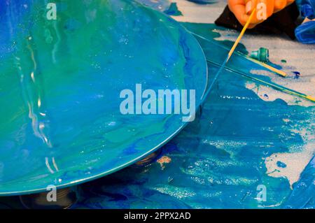 The process of creating a home-made trendy abstract modern pattern painted with a brush of acrylic blue multi-colored resin on a round wooden board. Stock Photo