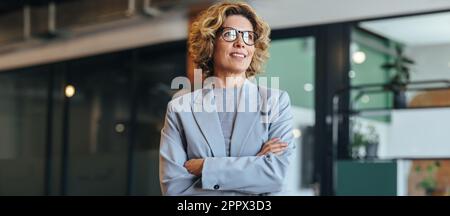 Thoughtful business woman standing with crossed arms. Woman in her 40's working in an office. Professional woman wearing a suit. Stock Photo