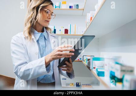 Doctor using a tablet while standing in a pharmacy. Female healthcare worker processing online prescriptions in a hospital dispensary. Woman working i Stock Photo