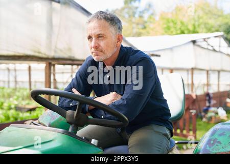 Thoughtful mature male farmer sitting with arms crossed on tractor by greenhouse Stock Photo