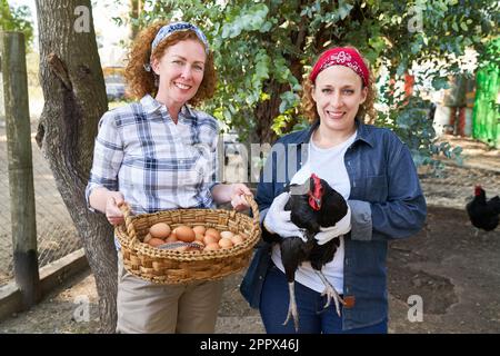 Portrait of smiling female farming coworkers holding black hen and wicker basket with brown eggs at poultry farm Stock Photo
