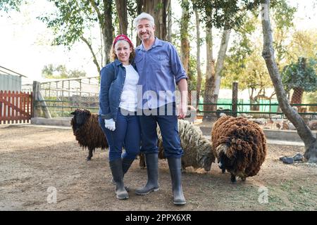 Full length portrait of smiling female and male farmers standing with sheep against trees at farm Stock Photo