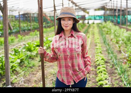 Portrait of smiling mature female farmer holding stick in organic farm on sunny day Stock Photo