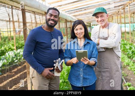 Portrait of smiling female farmer with tablet computer standing amidst male colleagues in greenhouse Stock Photo