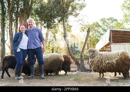 Full length portrait of happy male and female farmers standing with sheep against trees at farm Stock Photo