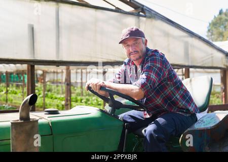 Portrait of mature male farmer sitting on tractor with greenhouse in background on sunny day Stock Photo