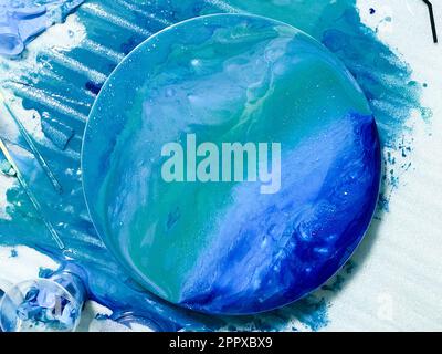 The process of creating a home-made trendy abstract modern pattern painted with a brush of acrylic blue multi-colored resin on a round wooden board. Stock Photo