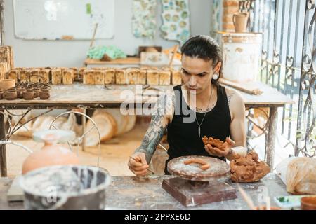 Concentrated young tattooed male potter in black tank top making clay product while working in professional workshop and creating earthenware Stock Photo