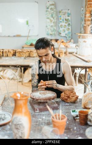 Focused young tattooed male artisan in black tank top working on clay wheel while shaping ceramic pot in professional workshop Stock Photo