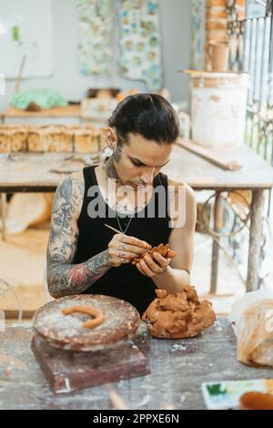 Concentrated young tattooed male potter in black tank top making clay product while working in professional workshop and creating earthenware Stock Photo