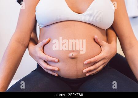 Crop specialist standing and treating pregnant patient sitting on table from behind while massaging tummy in clinic Stock Photo