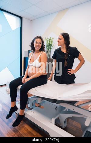 Full body of young pregnant female sitting on bench and looking away while enjoying therapy session of osteopath standing behind with hand on waist in Stock Photo