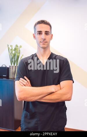 Confident young male in black medical scrubs with hands crossing chest standing near potted plant and looking at camera in light room Stock Photo
