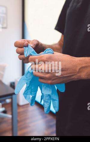 Unrecognizable male doctor in black t shirt putting on blue latex gloves while preparing for surgery in modern hospital Stock Photo
