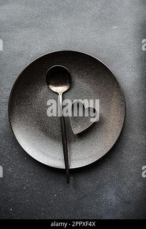 Minimalistic table setting in black color with plate and spoon on the same color table with heart on plate Stock Photo