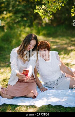 Calm teenage granddaughter sharing smartphone with grandmother while smiling and sitting on blanket together during picnic in park Stock Photo