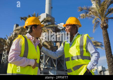 Happy young African American engineers in uniforms and helmets smiling while having conversation against building on sunny day Stock Photo