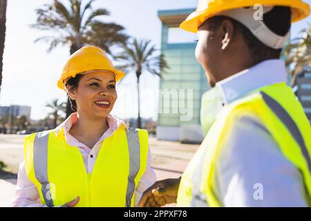 Happy young African American engineers in uniforms and helmets smiling while having conversation against building on sunny day Stock Photo