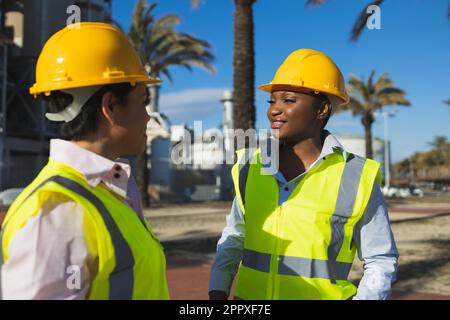 Happy young African American engineers in uniforms and helmets smiling while having conversation against building on sunny day Stock Photo