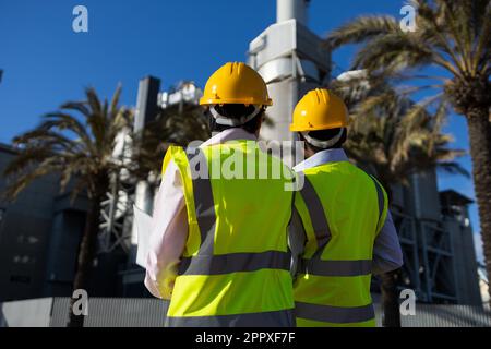 Back view of anonymous engineers in protective helmet and yellow vest standing together against building while working on construction Stock Photo
