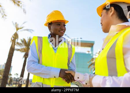 Side view of African American engineers in yellow waistcoat uniform happily shaking hands while standing looking at each other on city street over blu Stock Photo