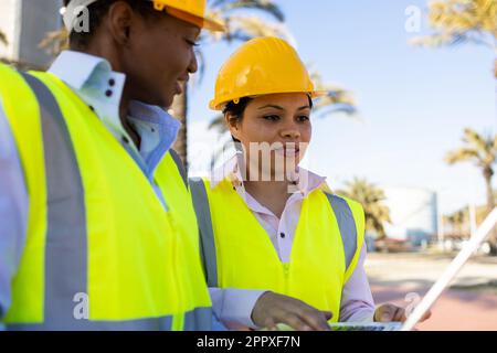 Builders in hardhat and engineer vest standing near construction site with laptop while discussing project Stock Photo