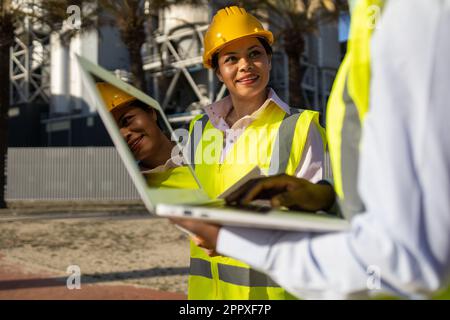 Happy young woman in yellow engineer uniform smiling and looking at colleague with laptop while working on construction site together Stock Photo